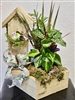 Rustic floral arrangement featuring a wooden birdhouse with moss accents, a colorful bird figurine, vibrant tropical greenery, and a decorative floral-patterned ribbon, all set in a natural wooden planter box.