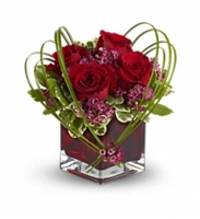 A romantic floral arrangement featuring lush red roses, pink waxflowers, and greenery, artistically designed in a red glass cube vase with decorative bear grass loops.