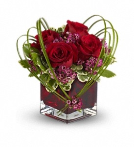 A romantic floral arrangement featuring lush red roses, pink waxflowers, and greenery, artistically designed in a red glass cube vase with decorative bear grass loops.