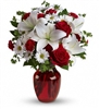 This image features a bouquet of red roses, white lilies, and gold-accented filler flowers arranged in a classic red glass vase.