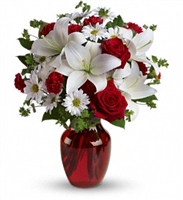 This image features a bouquet of red roses, white lilies, and gold-accented filler flowers arranged in a classic red glass vase.